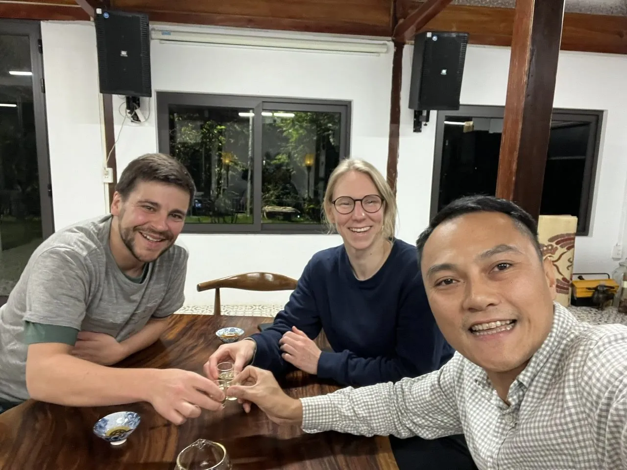 Three travelers and their guide clinking small glasses of corn wine at a wooden table