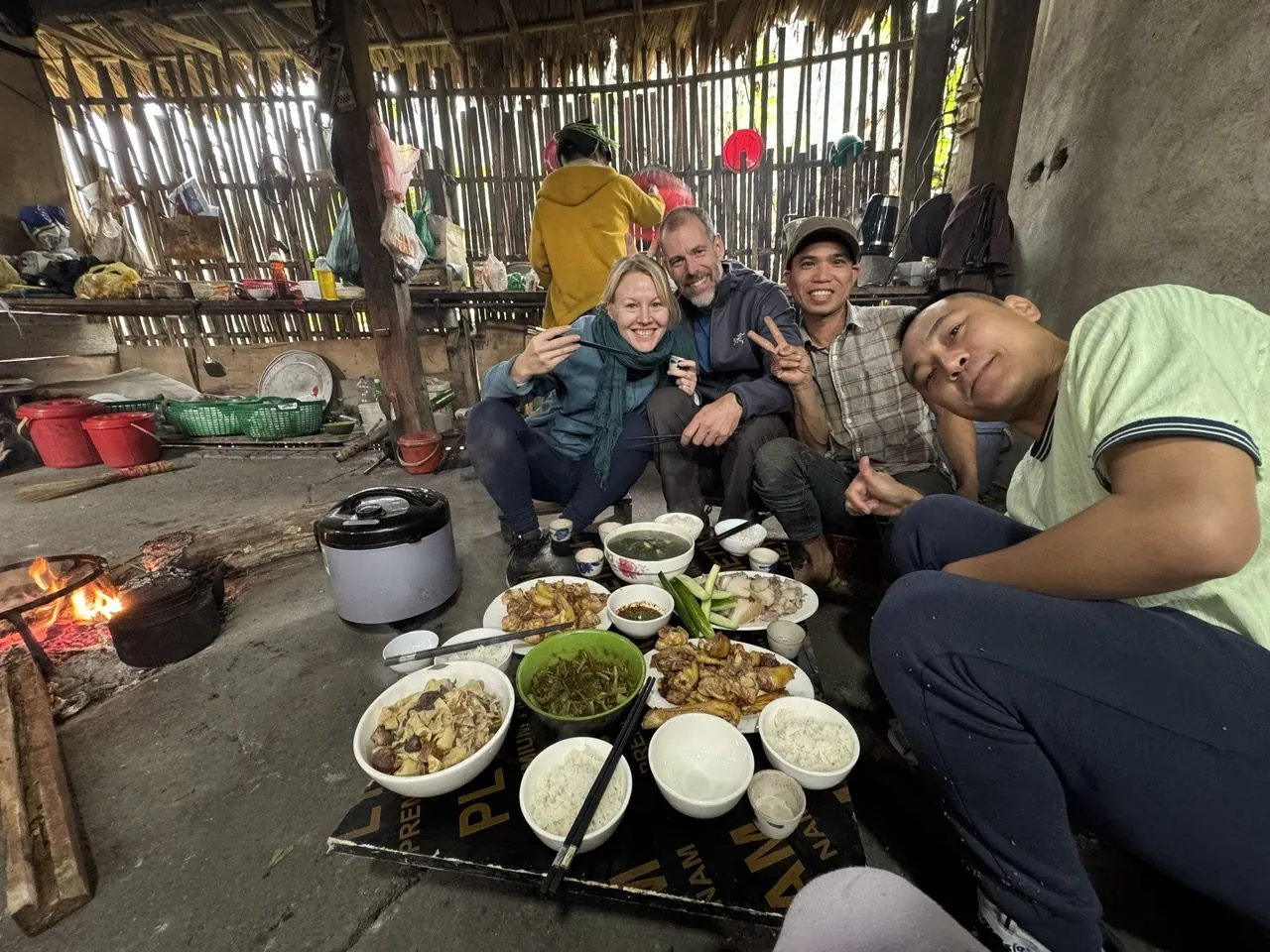 Travelers and their host sharing a home-cooked meal beside a cooking fire in a stilt-house kitchen